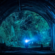 A shadowy figure stands at the opening of a dark tunnel, holding a flashlight.